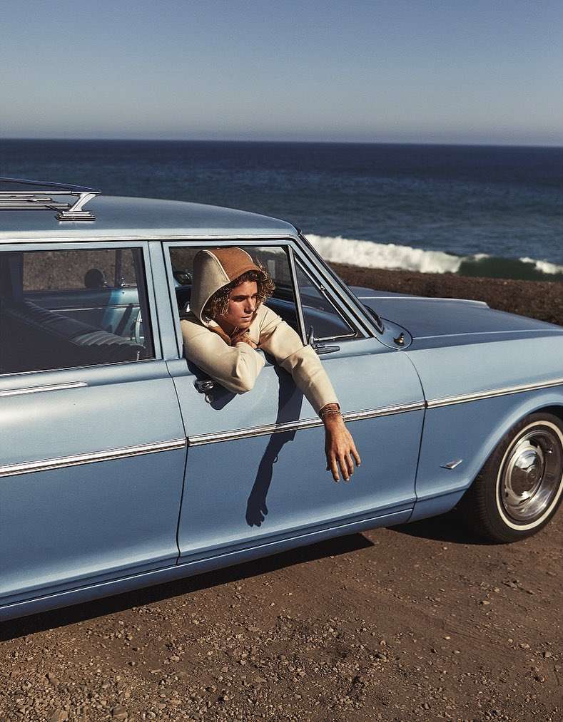 A young adult in hooded sweatshirt sits in a vintage blue car with the ocean and sky visible in the background.