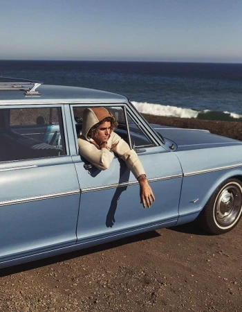 A young adult in hooded sweatshirt sits in a vintage blue car with the ocean and sky visible in the background.