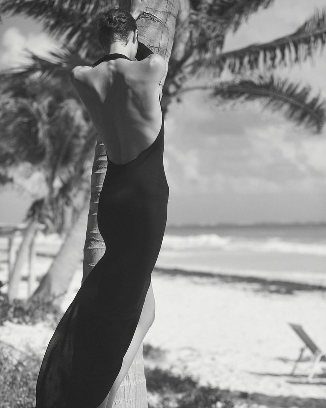 Model in a black backless dress leaning against a palm tree on a tropical beach in Los Angeles by Adam Franzino