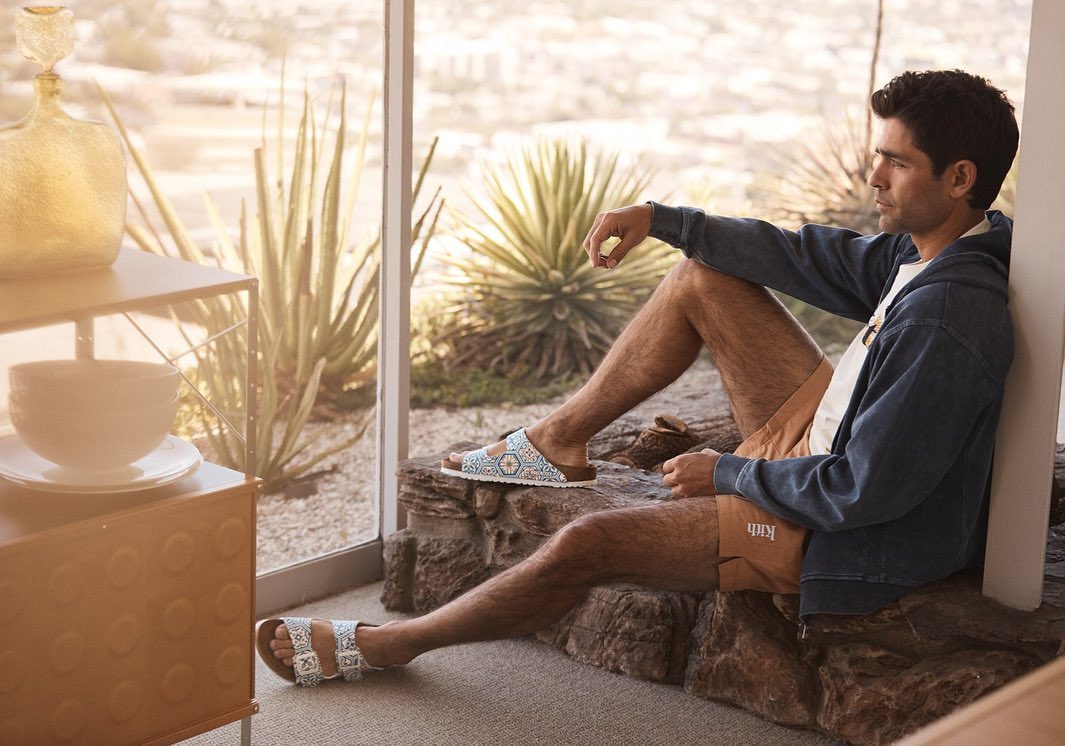 Man in blue hooded jacket and brown shorts sitting on rocks by window, wearing Birkenstock sandals outdoors in a desert setting.