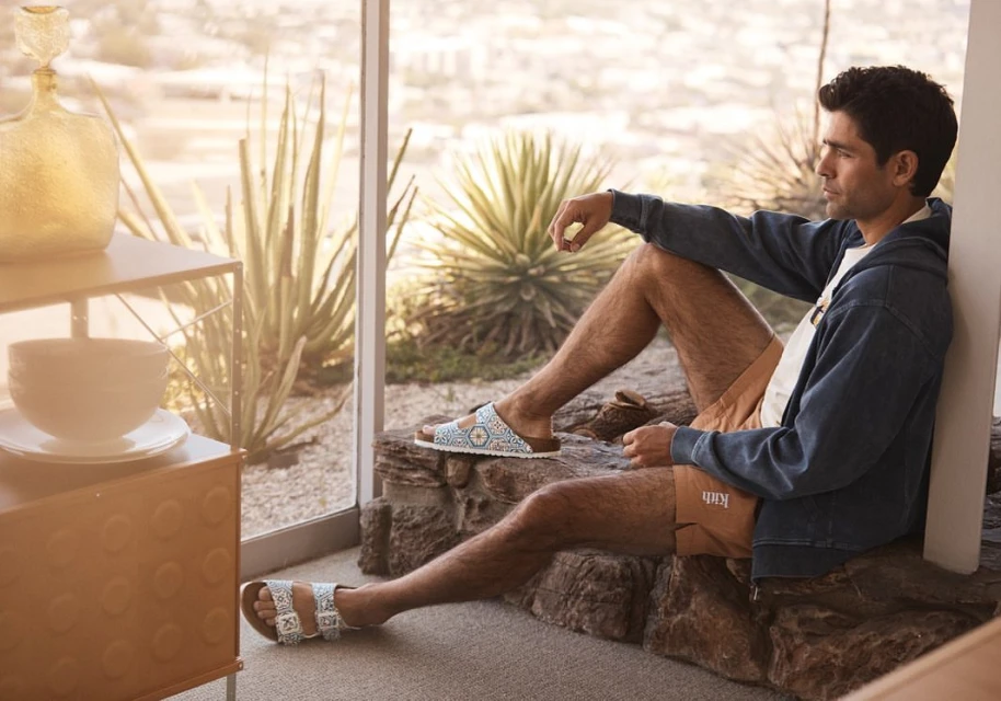 Man in blue hooded jacket and brown shorts sitting on rocks by window, wearing Birkenstock sandals outdoors in a desert setting.