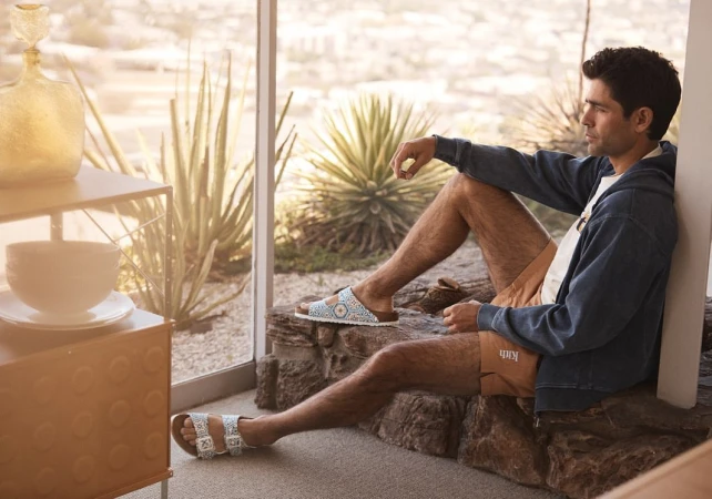 Man in blue hooded jacket and brown shorts sitting on rocks by window, wearing Birkenstock sandals outdoors in a desert setting.