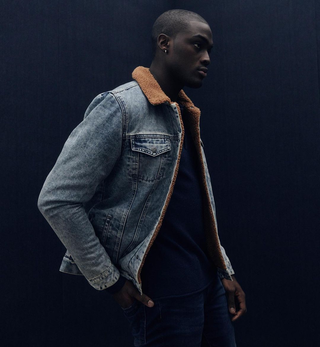 Dark-skinned adult male in a denim jacket with a fur collar and a navy shirt, standing against a dark background in a studio setting.