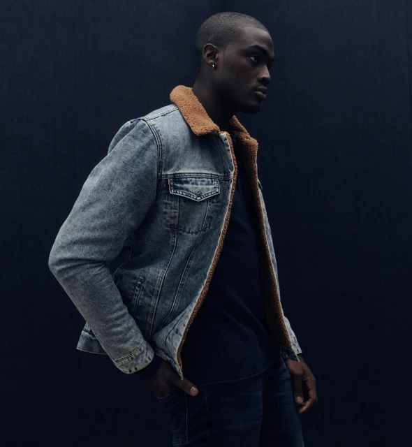 Dark-skinned adult male in a denim jacket with a fur collar and a navy shirt, standing against a dark background in a studio setting.