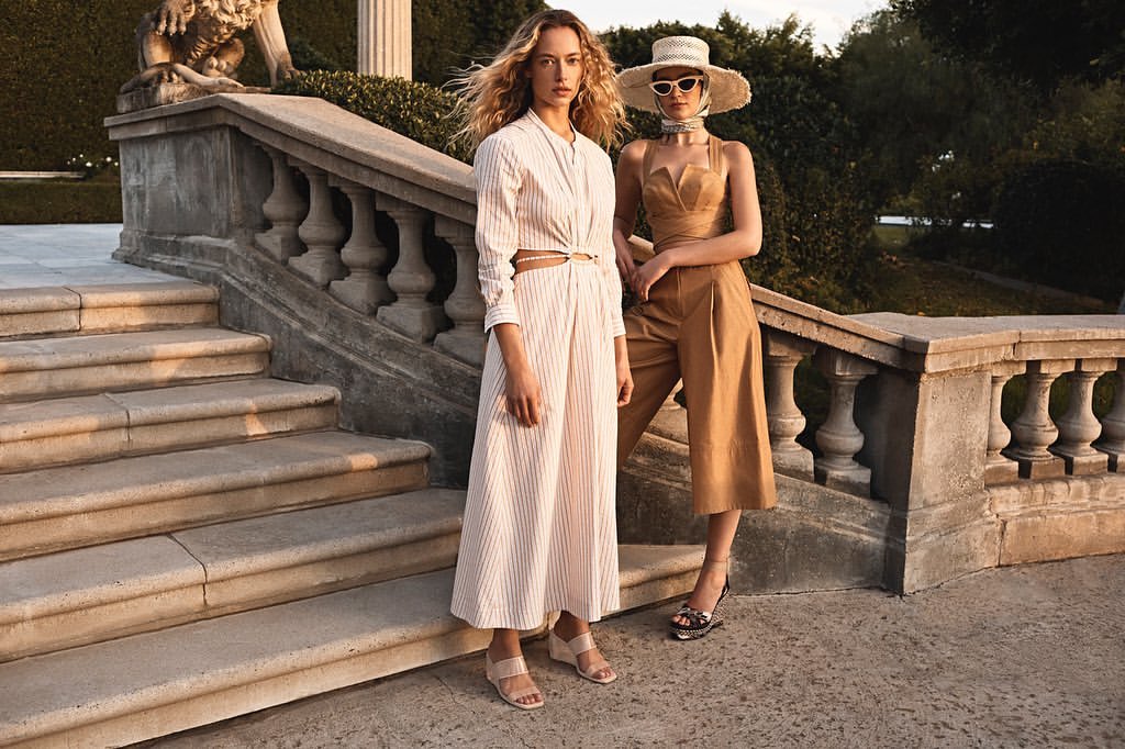 Two adult women pose on stone steps outdoors, showcasing neutral-toned fashion with a lion statue and green foliage in the background.