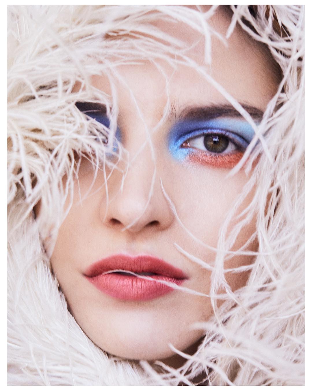 Close-up portrait of a woman with blue and orange eye makeup, framed by white feathers, capturing a beauty and fashion aesthetic.