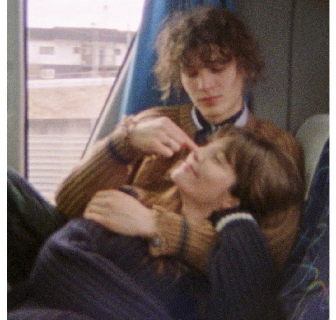 Image of a young couple embracing on a train, woman reclining against the man. He is touching her face, with a window in the background.
