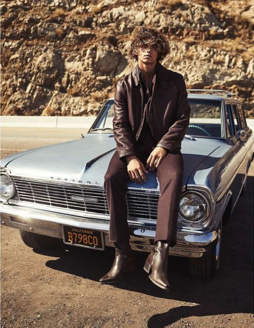 A person with curly hair sits on a light blue vintage car in a brown jacket and pants on a desert road in warm, natural lighting.