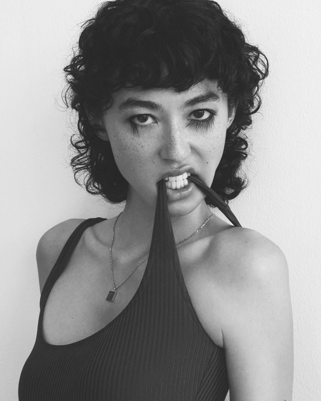 A striking black and white studio portrait of a young woman with short curly hair biting on a piece of licorice with dramatic makeup.