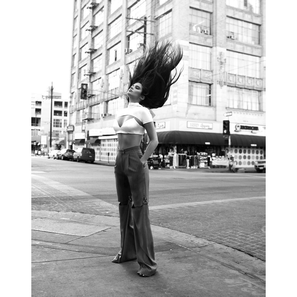 Monochrome portrait of a stylish adult woman with long dark hair tossing her head on a city sidewalk with tall buildings in the background.