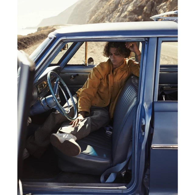 A young man sits inside a blue vintage car parked on a coastal road. He's wearing a yellow jacket and patterned pants, with a scenic view outside.