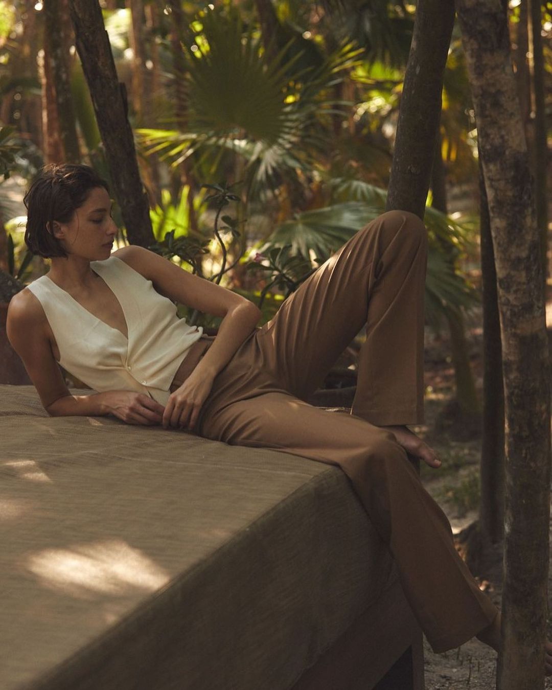 Woman in cream top and tan pants poses on surface in tropical forest with tall trees, dappled sunlight, and lush greenery in background.