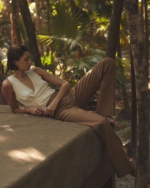 Woman in cream top and tan pants poses on surface in tropical forest with tall trees, dappled sunlight, and lush greenery in background.