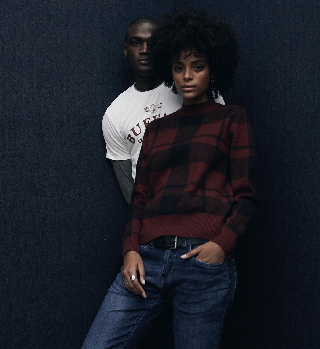 A woman in a plaid sweater and jeans poses with a man in a t-shirt in a fashion shot against a dark blue textured background.