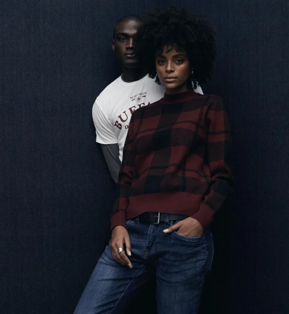 A woman in a plaid sweater and jeans poses with a man in a t-shirt in a fashion shot against a dark blue textured background.
