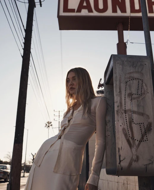 Adult female in a shirt and skirt leaning against a utility box near a laundromat sign on a city street at dusk or dawn.