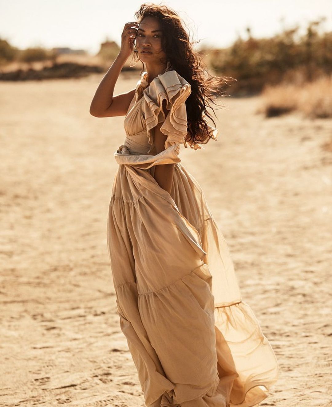 A woman in a beige dress poses in a desert setting, illuminated by soft, natural light, showcasing fashion and natural beauty.