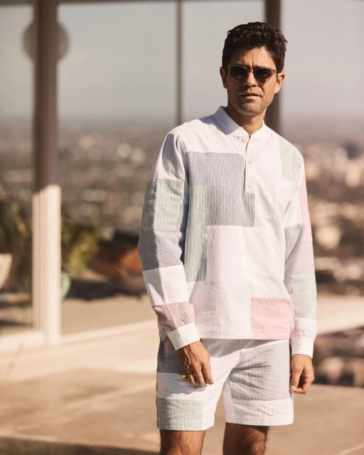 Man in a patchwork striped matching set outdoors on a sunny day. He's wearing sunglasses with a blurred city and sky background.