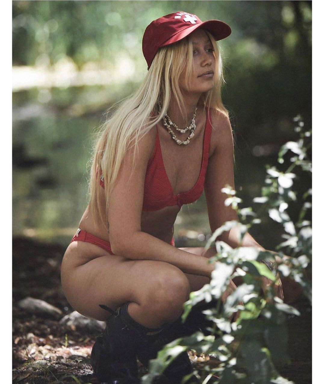 Blonde woman in a red bikini and baseball cap poses in a leafy, outdoor setting with a river. Fashion, swimwear, and nature.