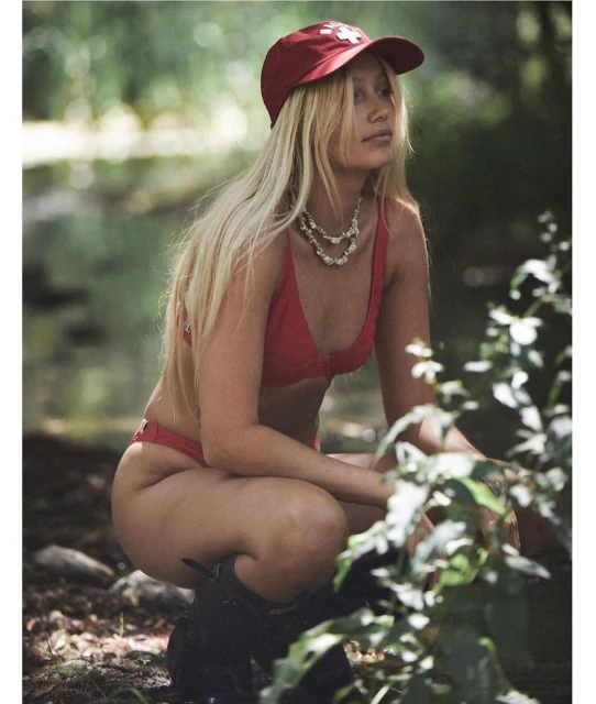 Blonde woman in a red bikini and baseball cap poses in a leafy, outdoor setting with a river. Fashion, swimwear, and nature.