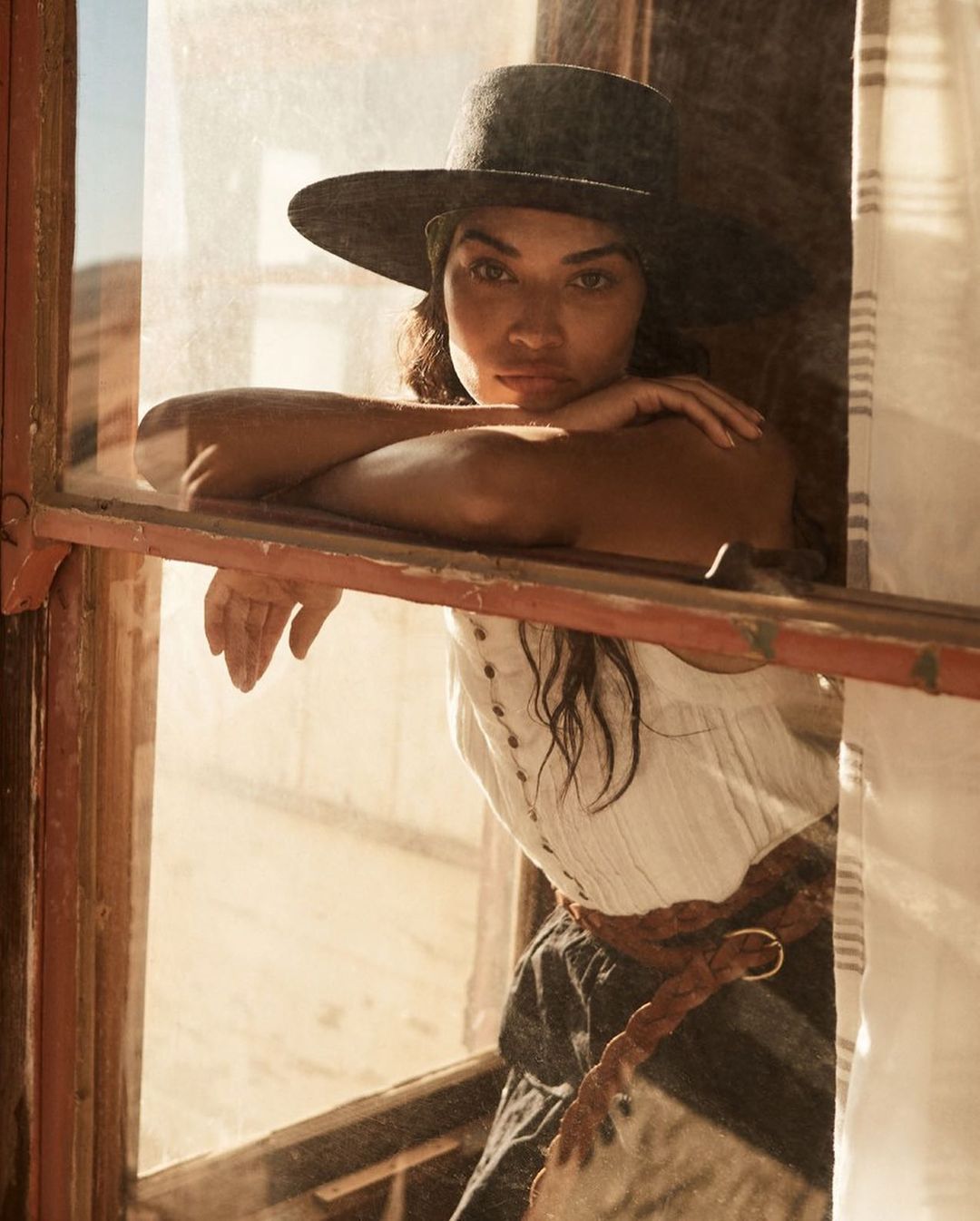 Portrait of a woman in a hat looking out a window in a sunlit desert setting, wearing casual clothing and a braided belt.