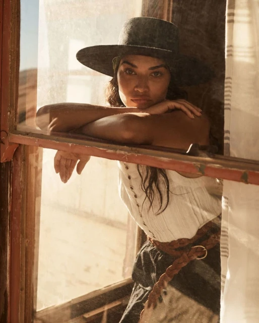 Portrait of a woman in a hat looking out a window in a sunlit desert setting, wearing casual clothing and a braided belt.