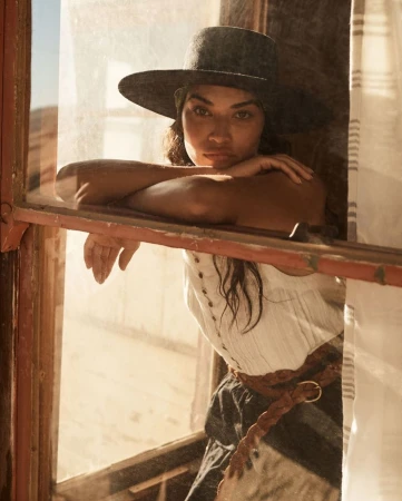 Portrait of a woman in a hat looking out a window in a sunlit desert setting, wearing casual clothing and a braided belt.