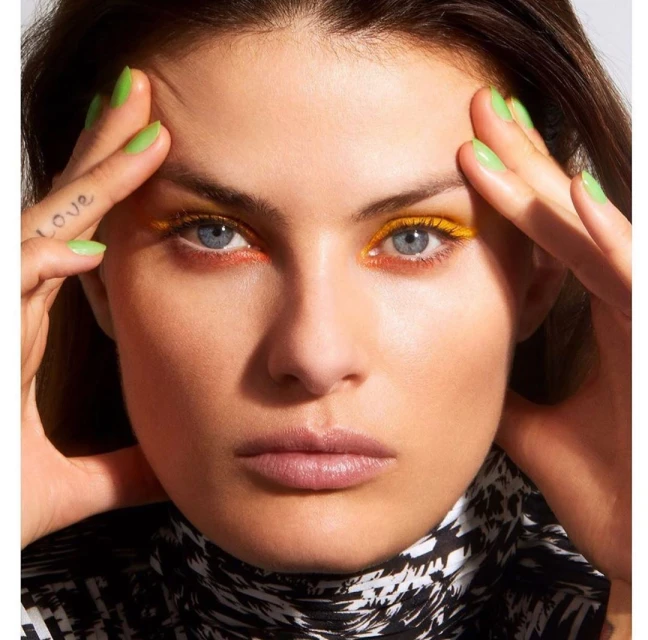 Close-up portrait of a woman with colorful eye makeup and green nail polish, hands framing face, wearing a black and white patterned turtleneck.