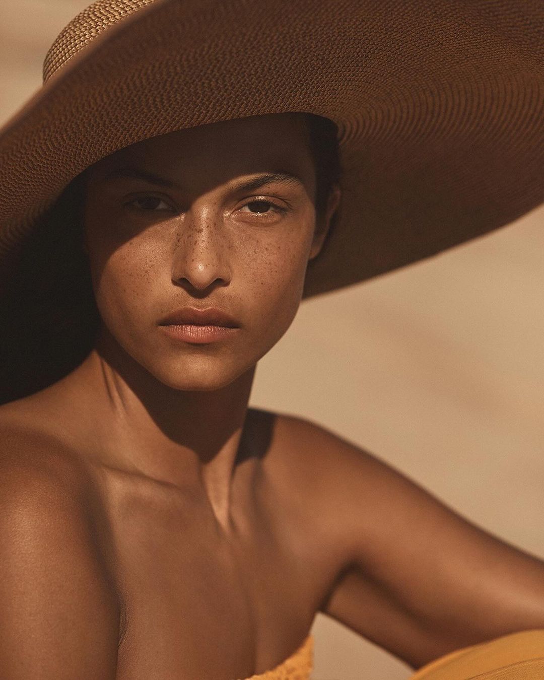 Close-up of an adult female with freckles, wearing a strapless top and a large straw hat, posing against a neutral background.