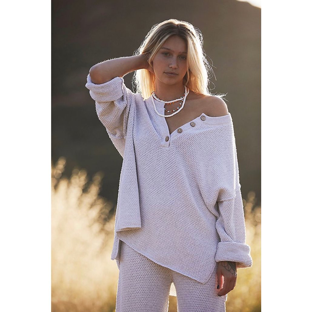 A young woman poses outdoors wearing a textured cream-colored sweater, matching pants, and beaded necklaces in soft, diffused light.