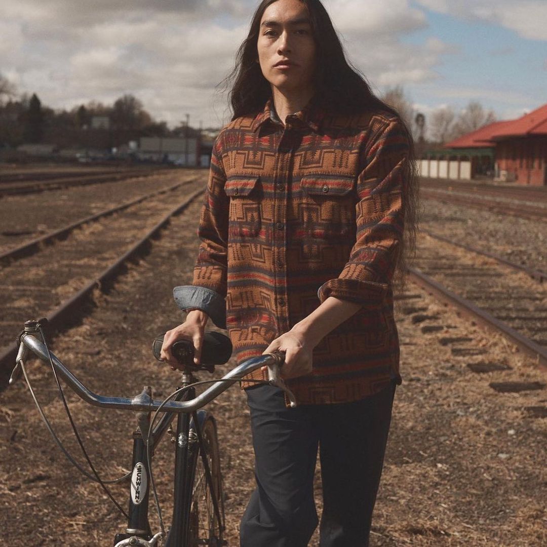 Young man with long hair stands with a vintage bicycle on railroad tracks in a flannel shirt with geometric patterns and black pants.