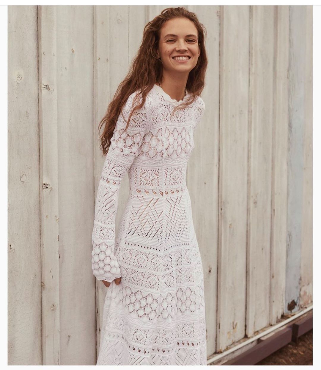A fair-skinned adult female with wavy brown hair smiles while wearing a long white lace dress, standing in front of a gray wooden wall.