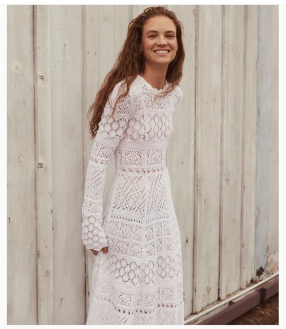 A fair-skinned adult female with wavy brown hair smiles while wearing a long white lace dress, standing in front of a gray wooden wall.