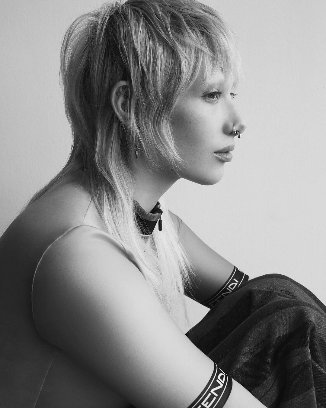 Monochrome portrait of a blonde woman with facial piercings sitting with her arms resting on her legs in an indoor setting against a white wall.