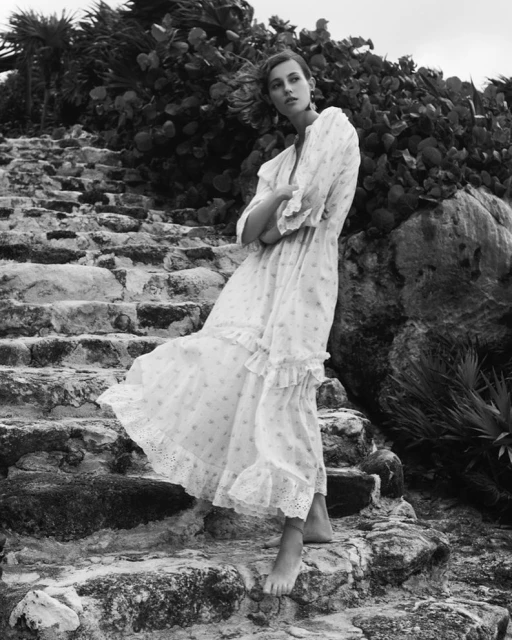 A woman in a white floral dress poses on stone steps outdoors, accented by lush vegetation, exuding a natural, serene vibe.