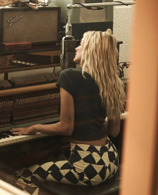 Blonde woman plays piano in a music studio, wearing patterned pants and a dark t-shirt, with a vintage microphone and amp visible.