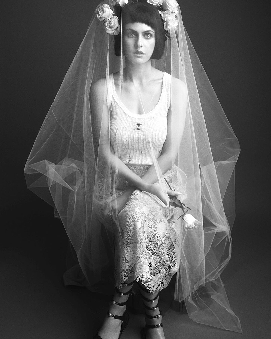 Monochrome portrait of a woman in a tank top, lace skirt, and gladiator sandals, wearing a veil and flower crown against a dark gray backdrop.