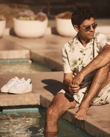 An adult male wearing a floral shirt and shorts sits with his feet in a pool, while white sneakers sit beside him on the pool ledge.