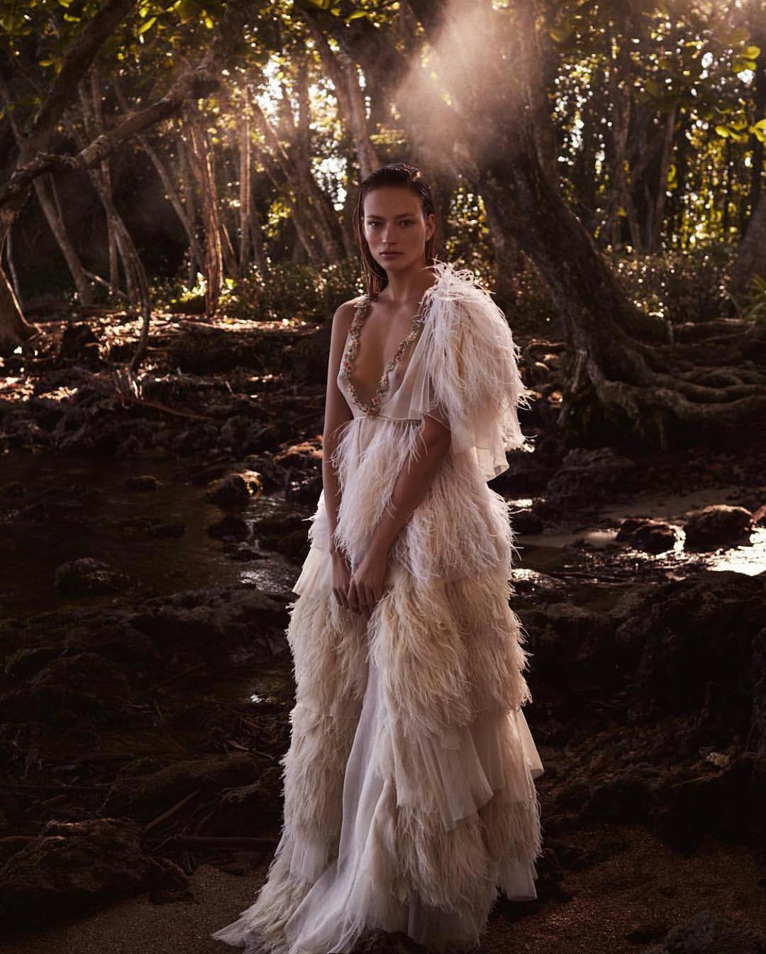 Adult female wearing a feather gown in a forest, shot with natural lighting. She stands in a stream surrounded by trees.