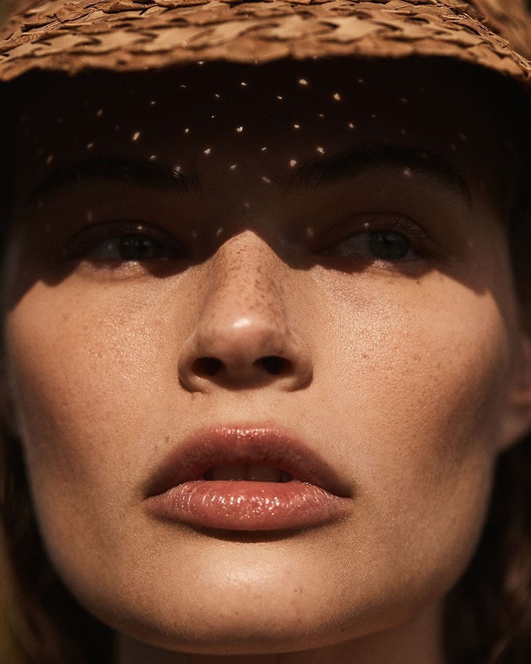 Close-up of a woman's face in dappled sunlight, wearing a straw hat casting shadows across her eyes and forehead, focusing on natural beauty.