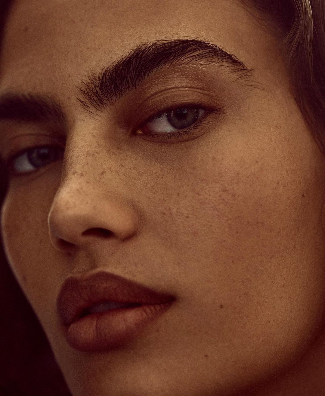 Close-up portrait of an adult woman with warm skin tones, prominent freckles, dark eyebrows, and brown lips.