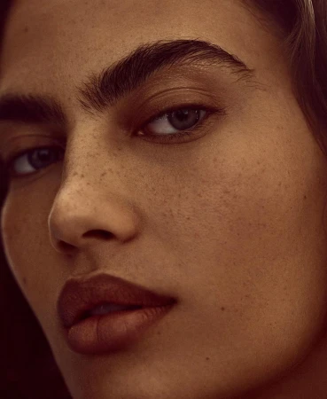 Close-up portrait of an adult woman with warm skin tones, prominent freckles, dark eyebrows, and brown lips.