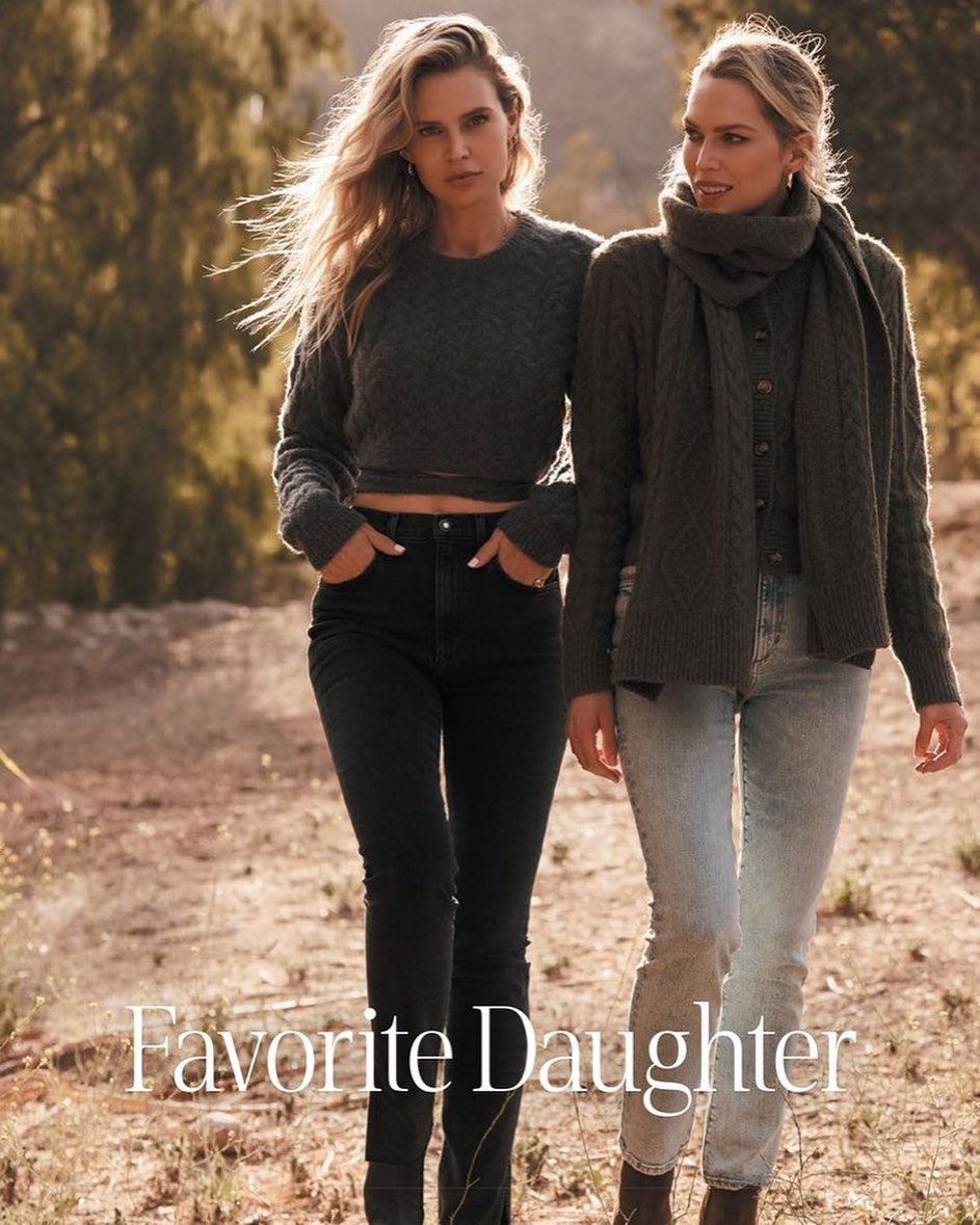Two adult females pose outdoors in casual grey knitwear and denim, standing on a dirt path with a natural background of trees and foliage.