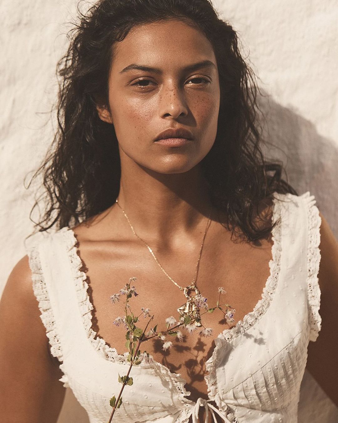 Portrait of a woman with dark hair and freckles wearing a white top and gold necklace, holding a sprig of flowers against a white wall.