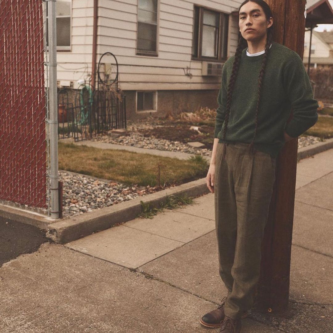Adult male with long braids poses outdoors in a green sweater and brown pants against a house with a street and fence.
