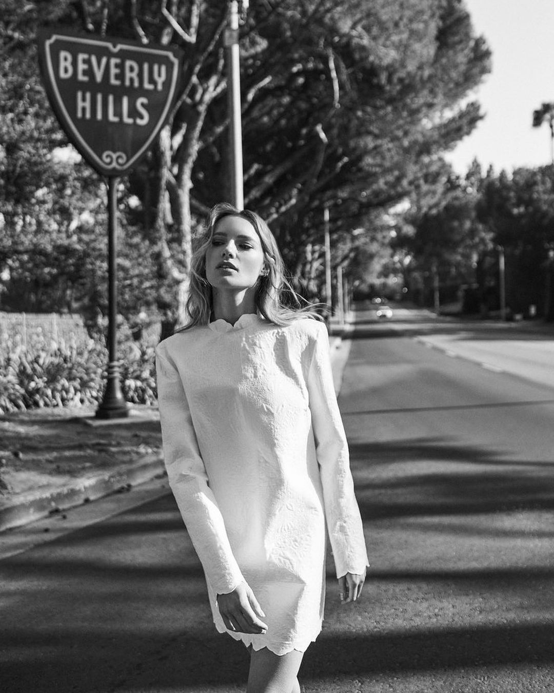 Black and white shot of a woman in a dress standing next to a Beverly Hills sign on a city street with trees and houses in the background.