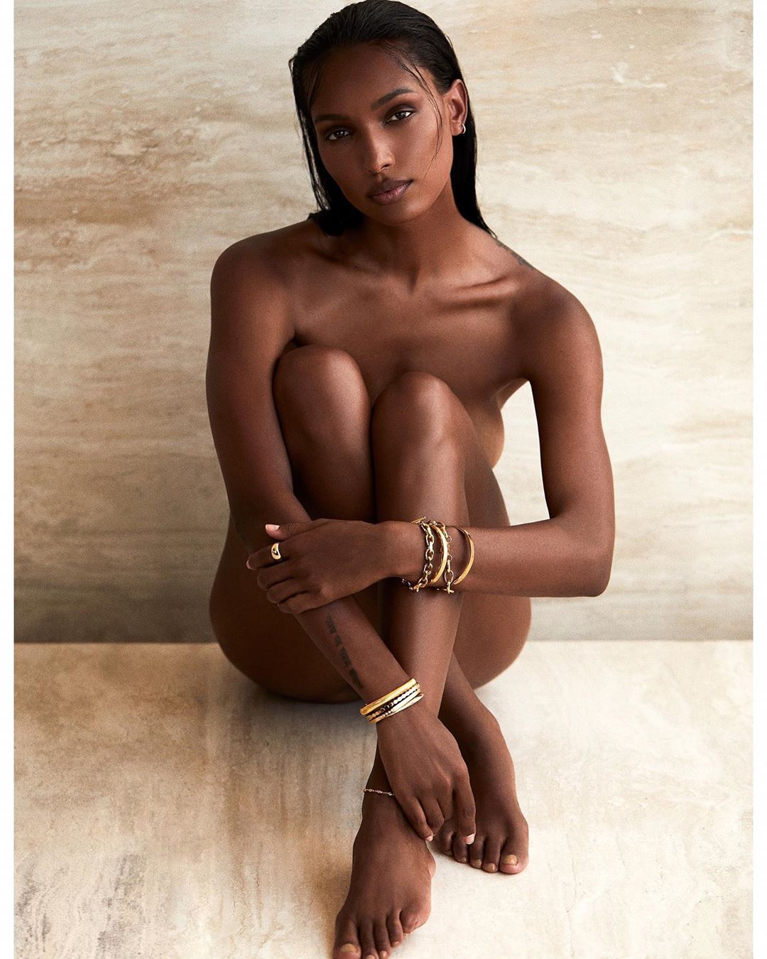 Glamorous portrait of a nude adult woman with dark skin, seated indoors against a wall, wearing bracelet jewelry, with arms around legs.