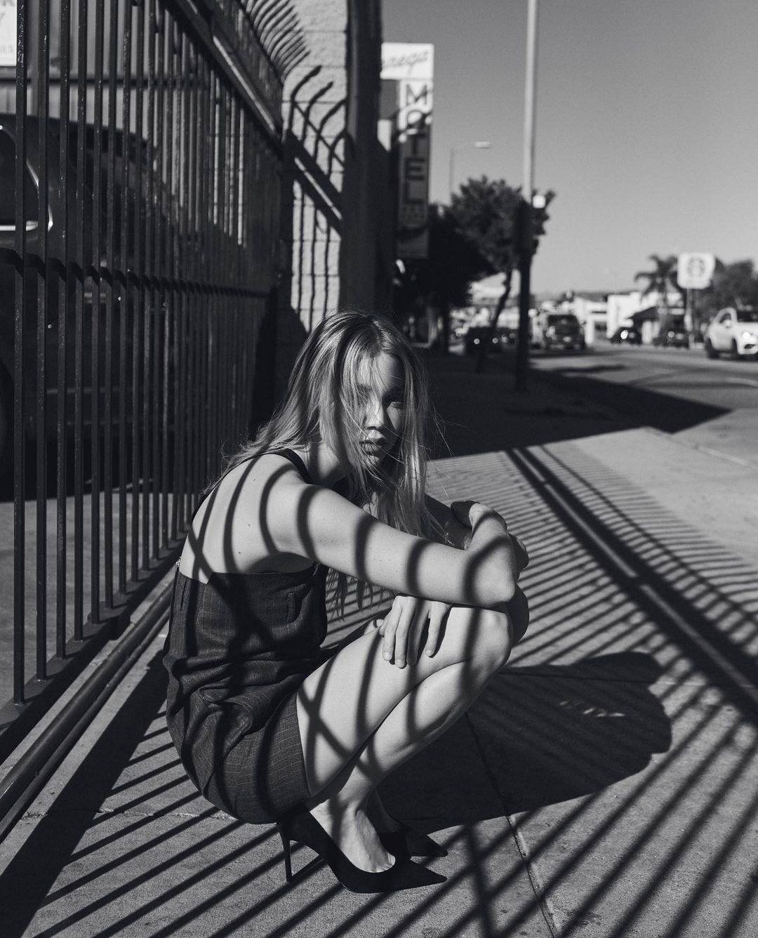 Black and white photograph of a blond woman in a dress and heels crouching behind a metal fence on a city sidewalk.