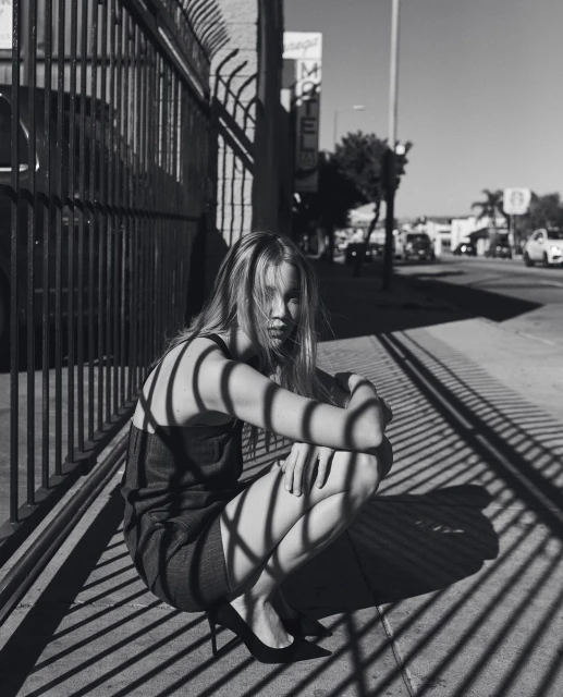 Black and white photograph of a blond woman in a dress and heels crouching behind a metal fence on a city sidewalk.
