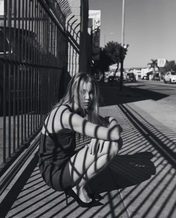 Black and white photograph of a blond woman in a dress and heels crouching behind a metal fence on a city sidewalk.